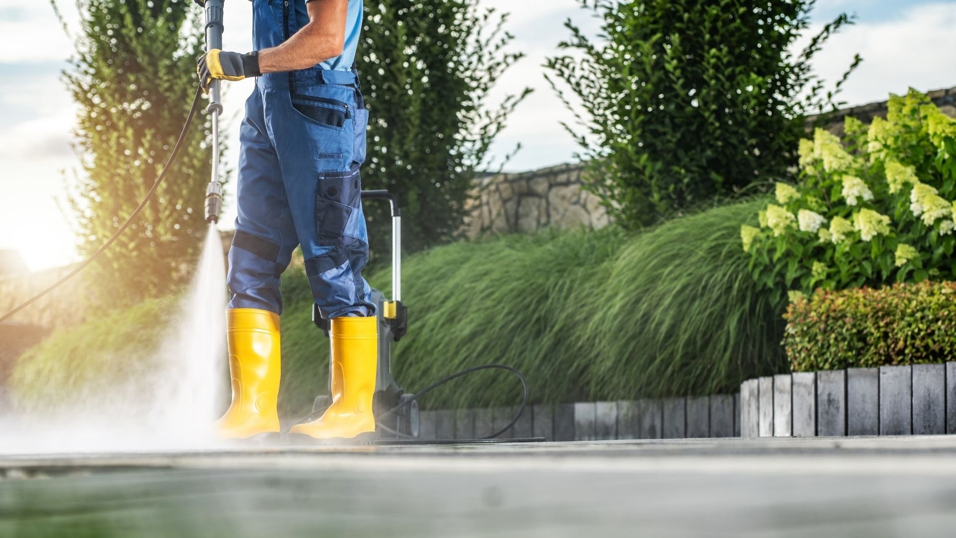 Worker in blue overalls and yellow boots pressure washing surface near hedges