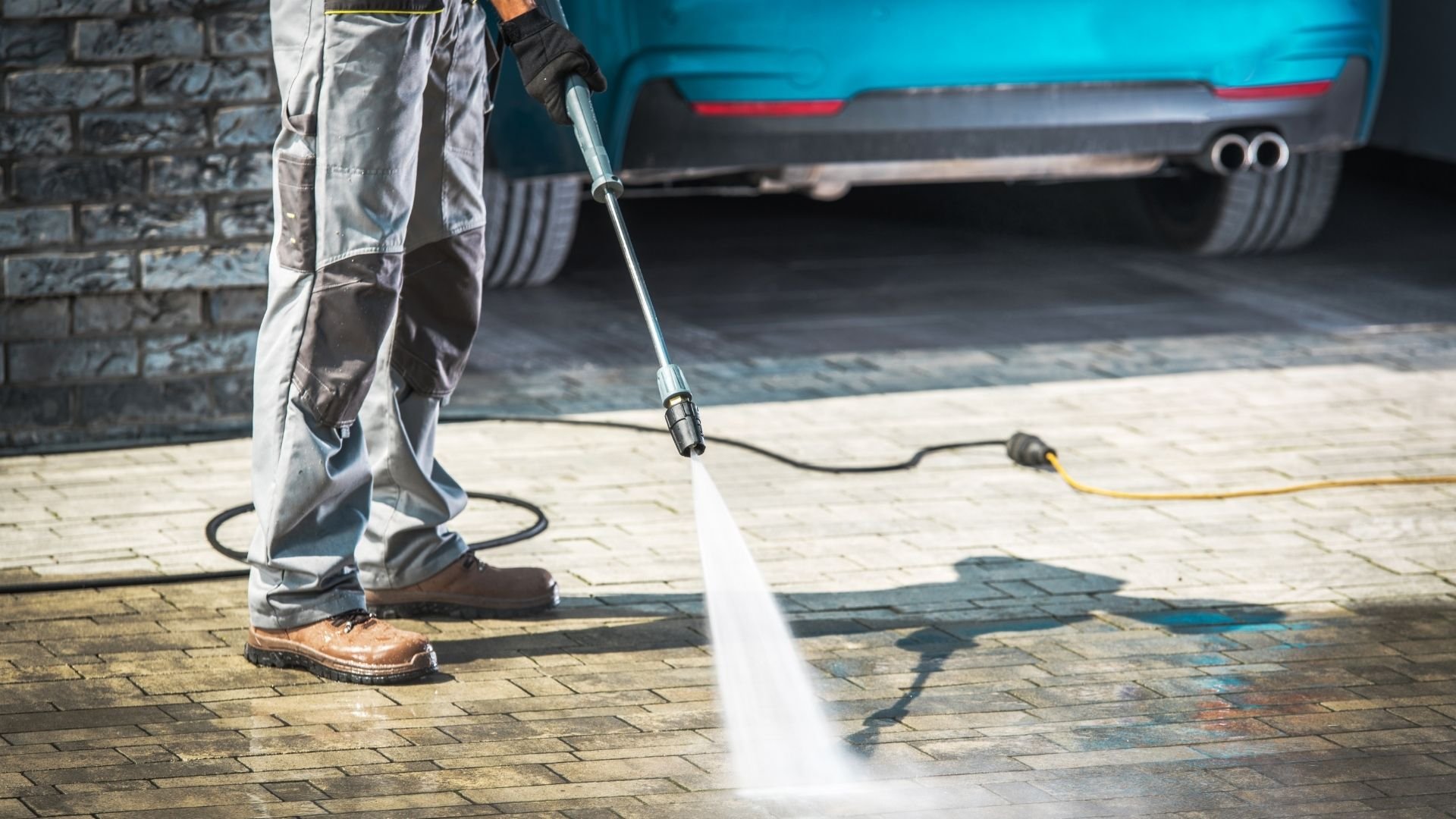 Worker pressure washing driveway with high-pressure spray hose near blue car